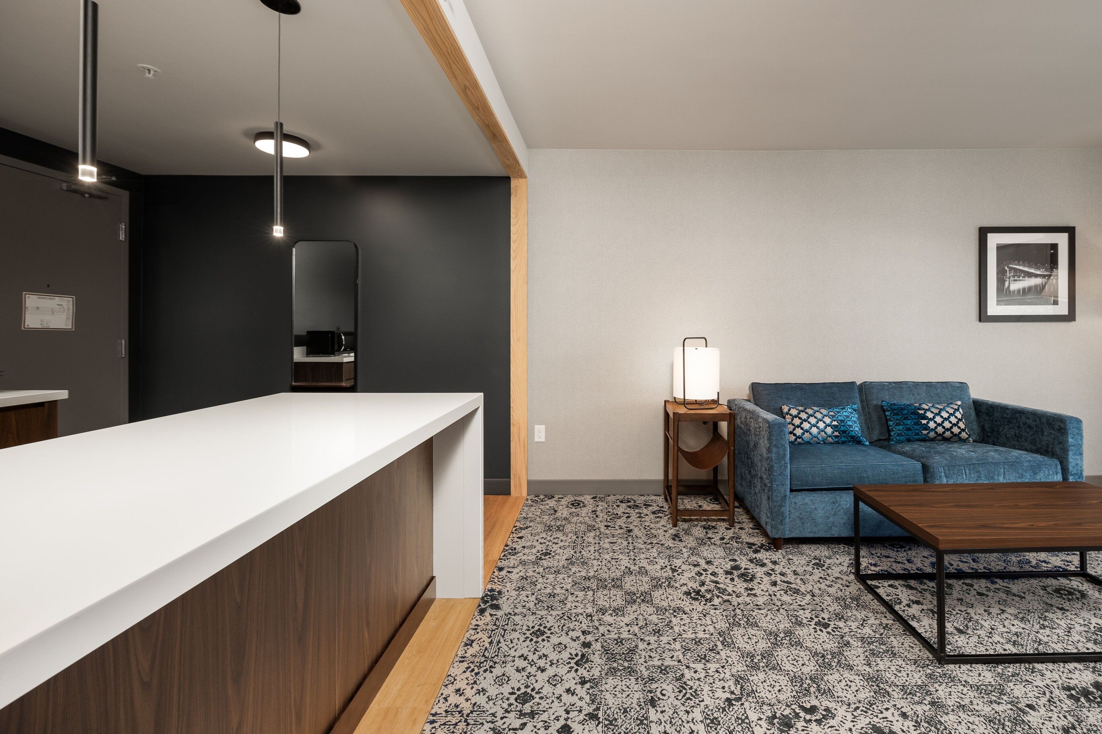 A modern kitchen with a white countertop, a wooden cabinet, and a black backsplash. There is a blue sofa with patterned cushions and a wooden coffee table. A small table with a lamp is also visible.
