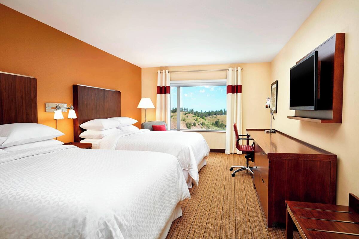 Quarto de hotel com duas camas, uma escrivaninha, uma cadeira, televisão e janela com vista para o exterior.
