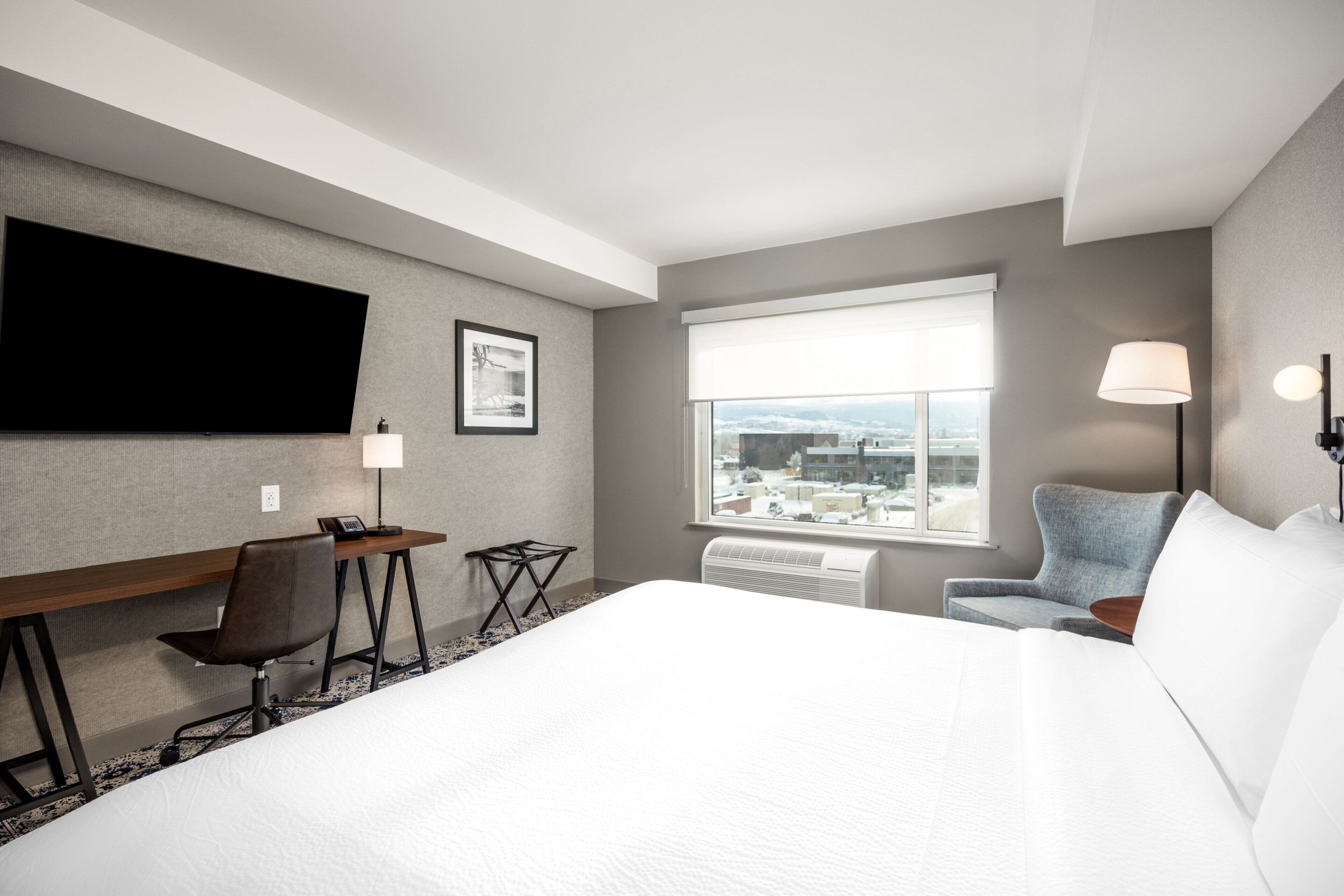 Une chambre d’hôtel avec un grand lit, un bureau avec une chaise, un téléviseur à écran plat, une fenêtre donnant sur la ville et une lampe fixée au mur.