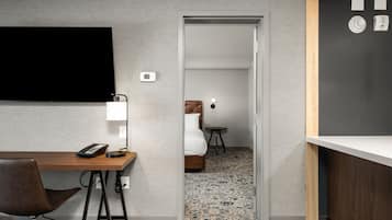 A hotel room with a wooden desk, a chair, a bed, a small table, a TV mounted on the wall, and a door leading to another room.