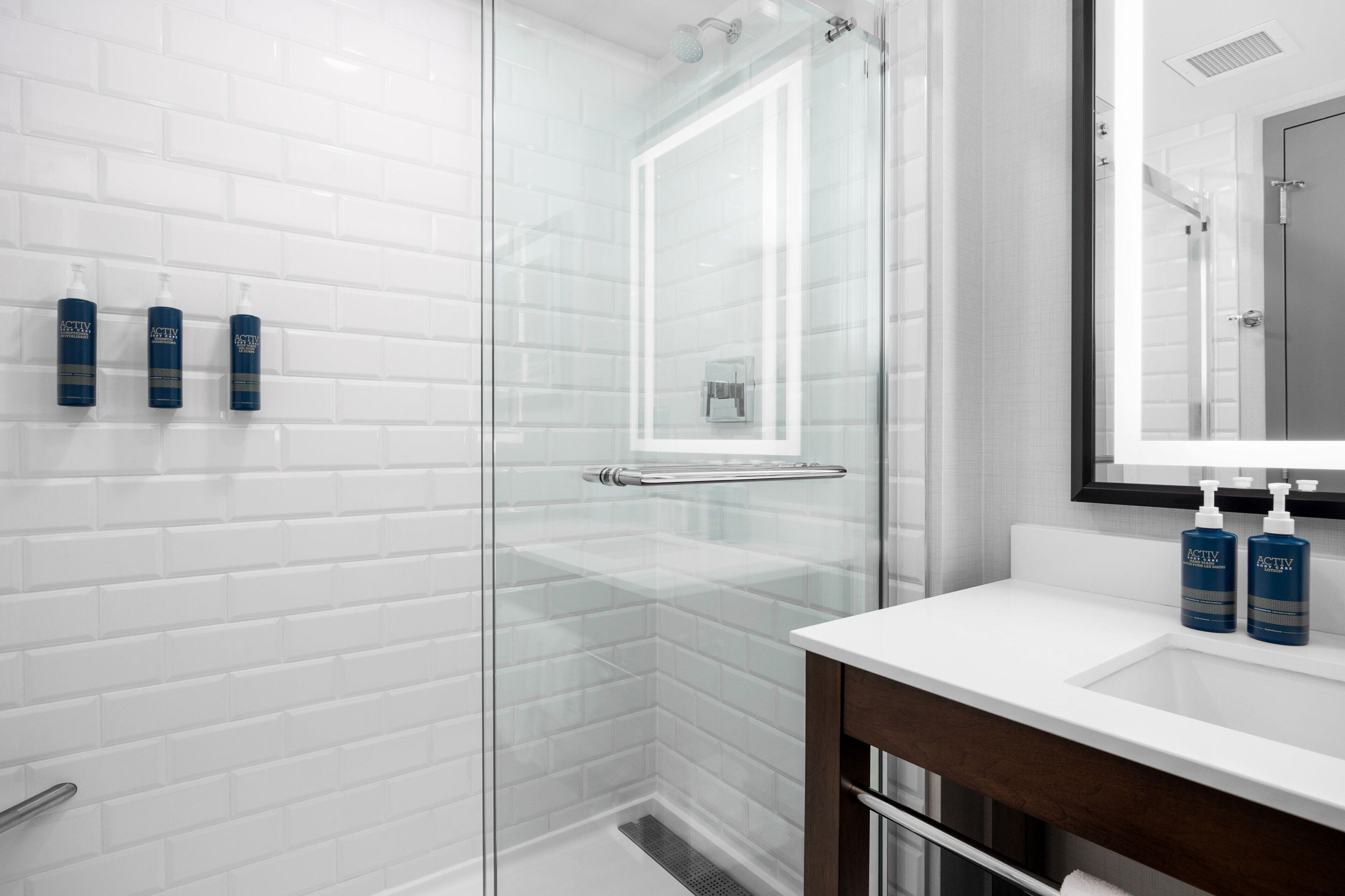 A modern bathroom with white subway tiles, a glass shower enclosure, and a white sink with two bottles of ACTIV soap.