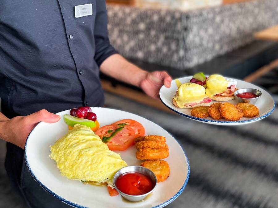 A person holding two plates with food items including eggs benedict, croquettes, and vegetables.