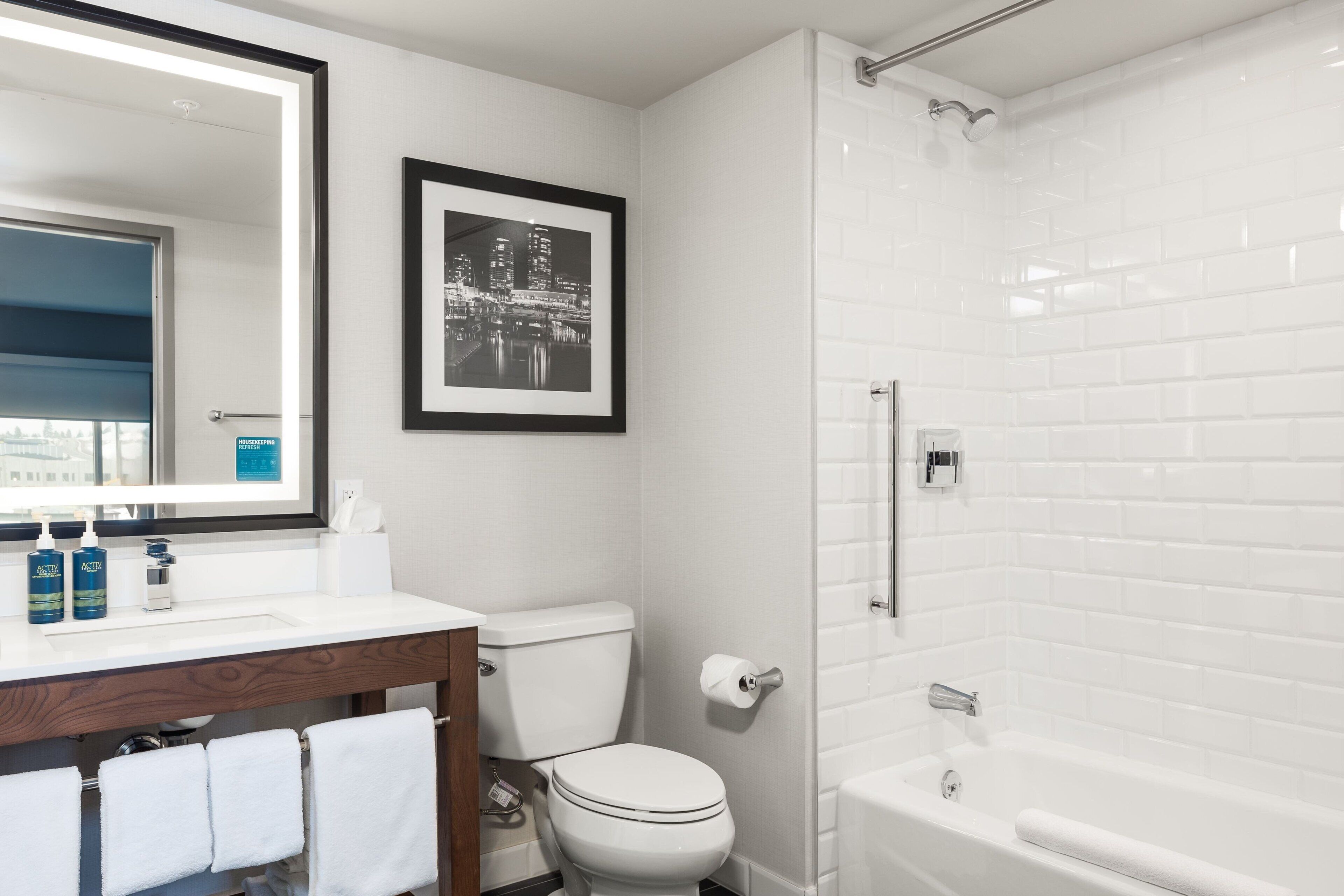 A bathroom with a white toilet, a sink with a mirror, a framed picture on the wall, and a white bathtub.