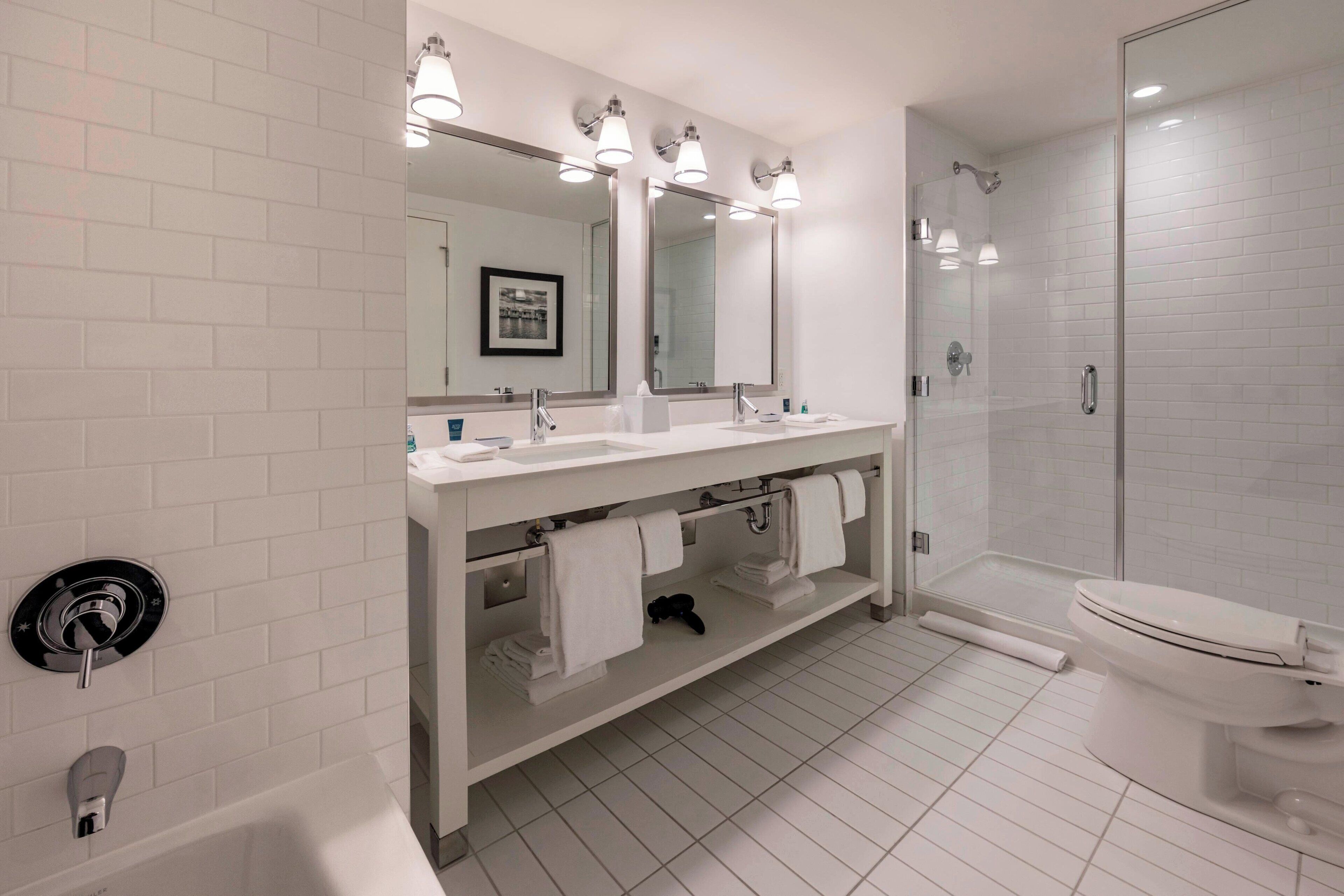 A modern bathroom with white tiled walls, a double sink vanity, a large mirror, a glass-enclosed shower, and a toilet.