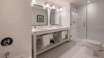 A modern bathroom with white tiled walls, a double sink vanity, a large mirror, a glass-enclosed shower, and a toilet.