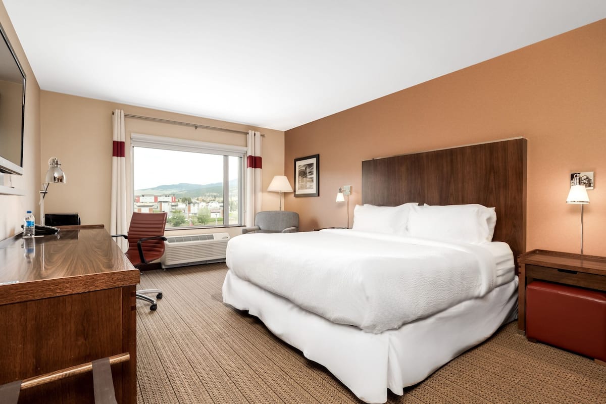 Quarto de hotel com uma cama grande, uma escrivaninha, uma cadeira, janela com vista e cabeceira de madeira.