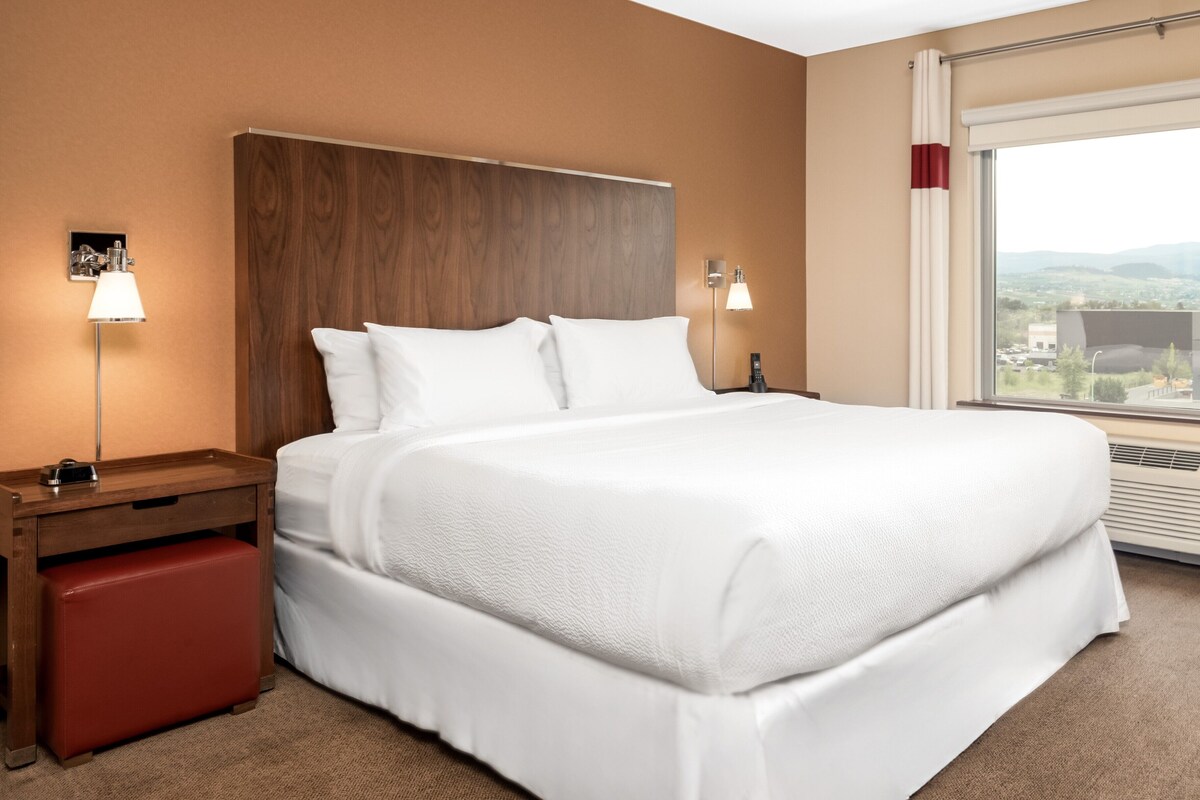 Habitación de hotel con una cama grande, cabecera de madera, lámparas de noche, un banco rojo y un amplio ventanal con vistas a la ciudad.
