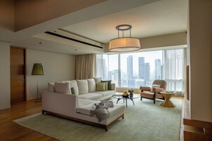 Una sala de estar moderna con sofá, sillón y mesa de centro, ventanas amplias con vistas a la ciudad y una lámpara colgante.