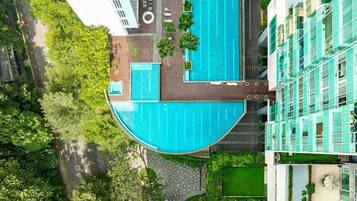 Oversigtsbillede af et moderne hotel med en stor swimmingpool, omgivet af frodig vegetation og beliggende ved en vej.