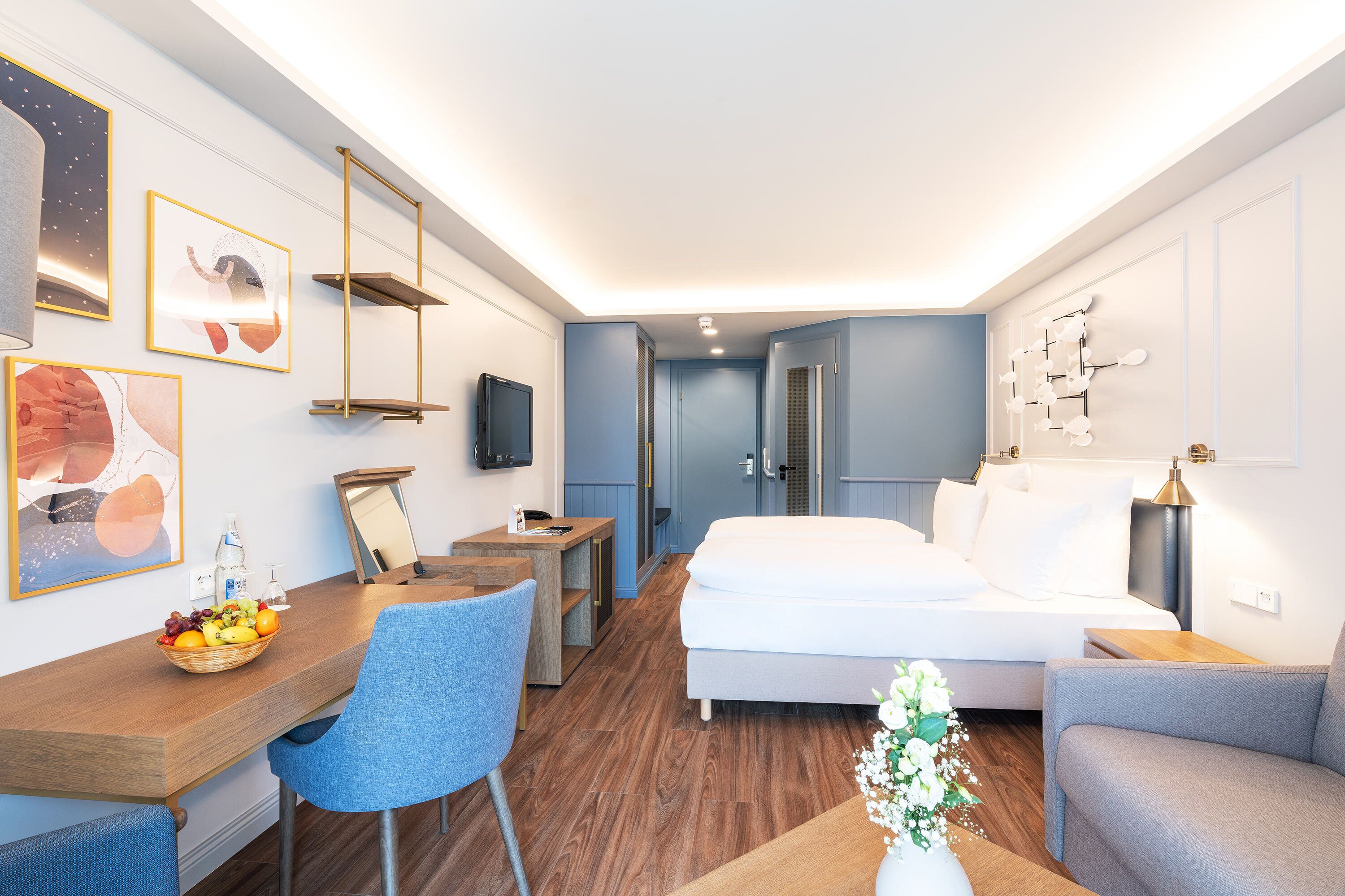 A modern hotel room with a bed, a desk with a chair, a small table with a vase, and artwork on the walls.