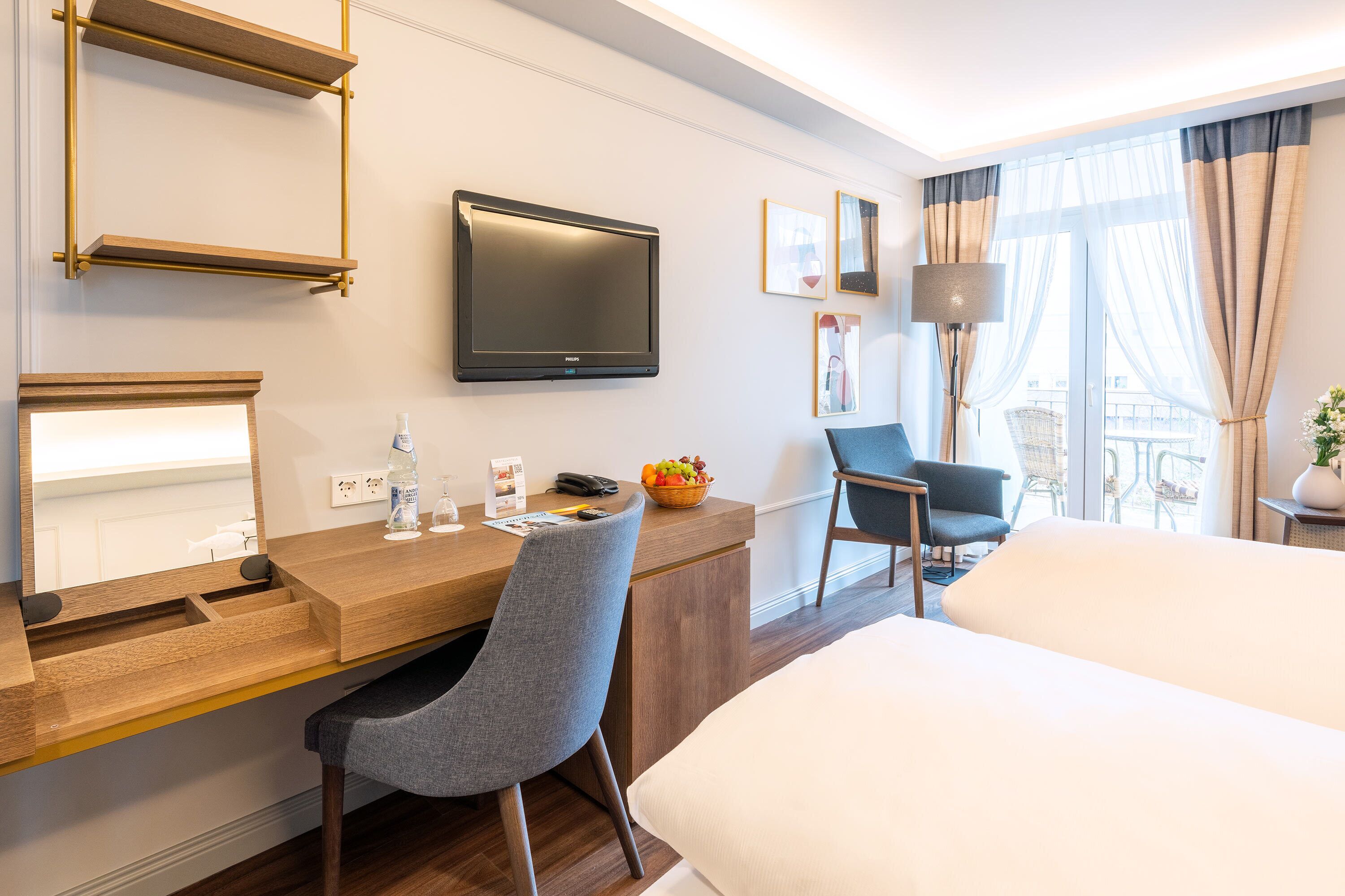 A hotel room with a wooden desk, a flat-screen TV, a comfortable chair, a bed, and a window with curtains.