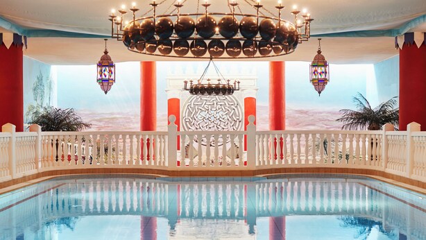 Une grande piscine intérieure agrémentée d’une balustrade décorative, d’un lustre et d’une fresque représentant un paysage désertique.
