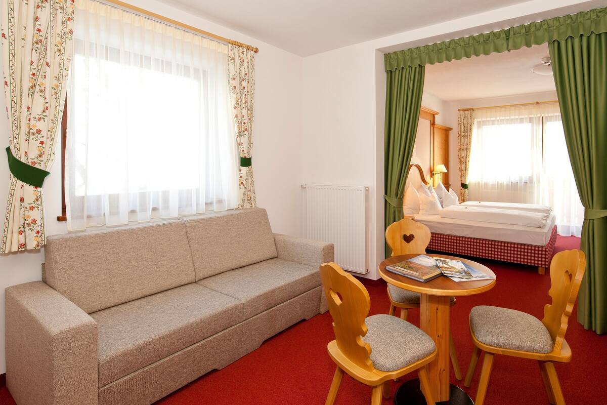 A hotel room with a sofa, two wooden chairs, a small table with magazines, a bed with a checkered bedspread, and green curtains.