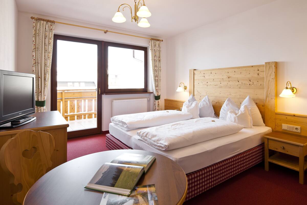 A hotel room with a bed, a desk with a computer, a window with curtains, and a wooden headboard.