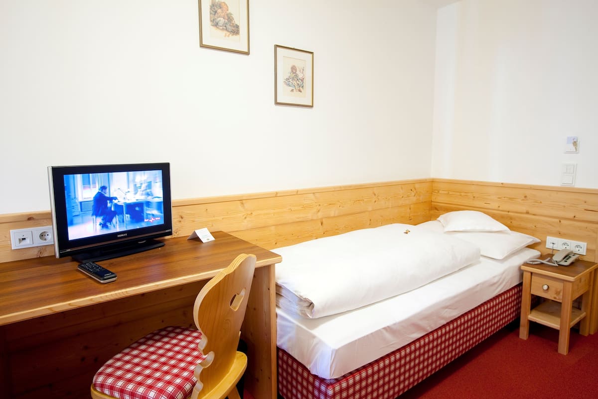 A hotel room with a wooden desk, a television, a bed with white linens, a chair, and a small bedside table with a phone.