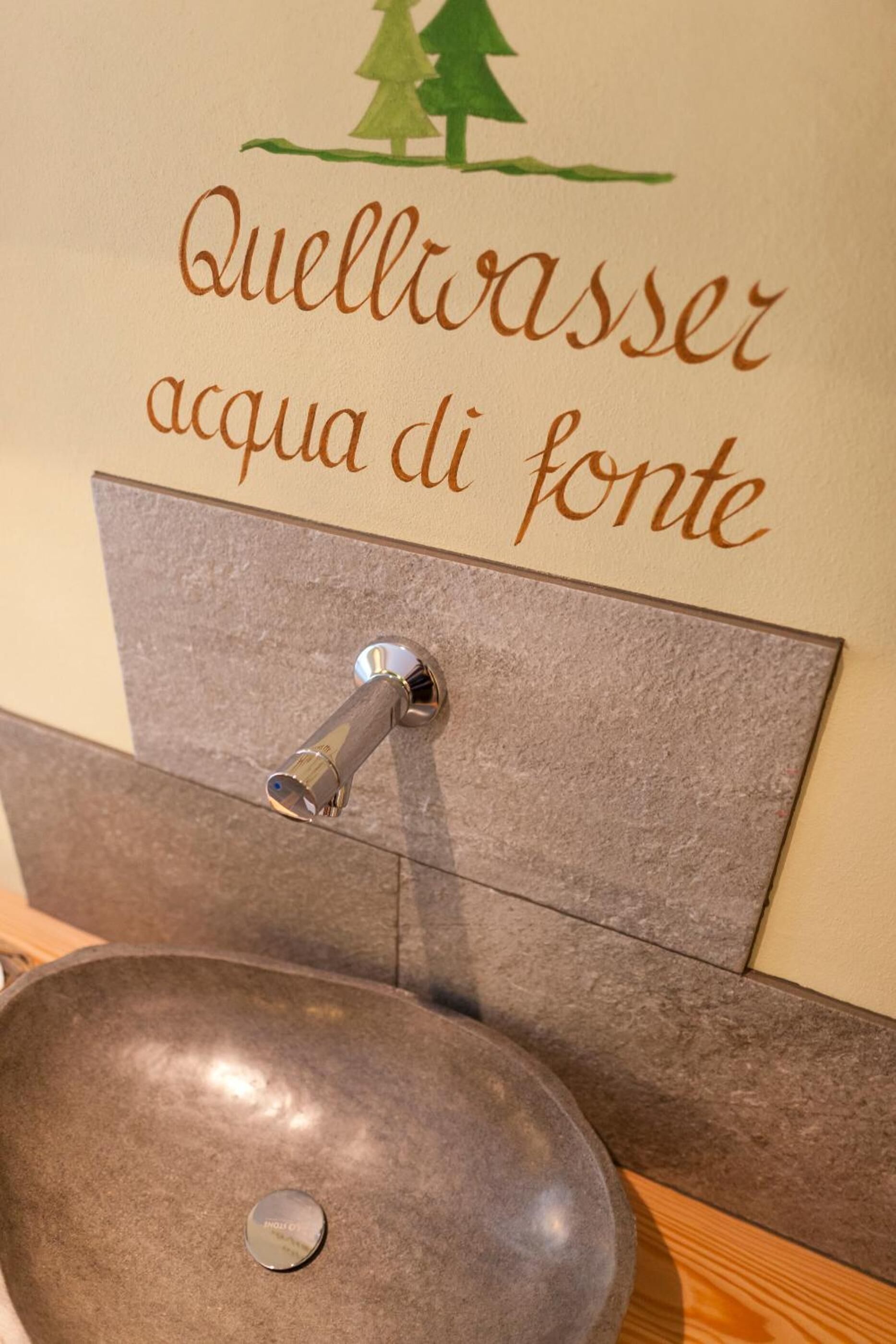 Un lavamanos con una pileta de piedra y un grifo de pared. Sobre el lavamanos, hay un letrero con el texto "Quellwasser acqua di fonte" y una pequeña ilustración de un árbol.
