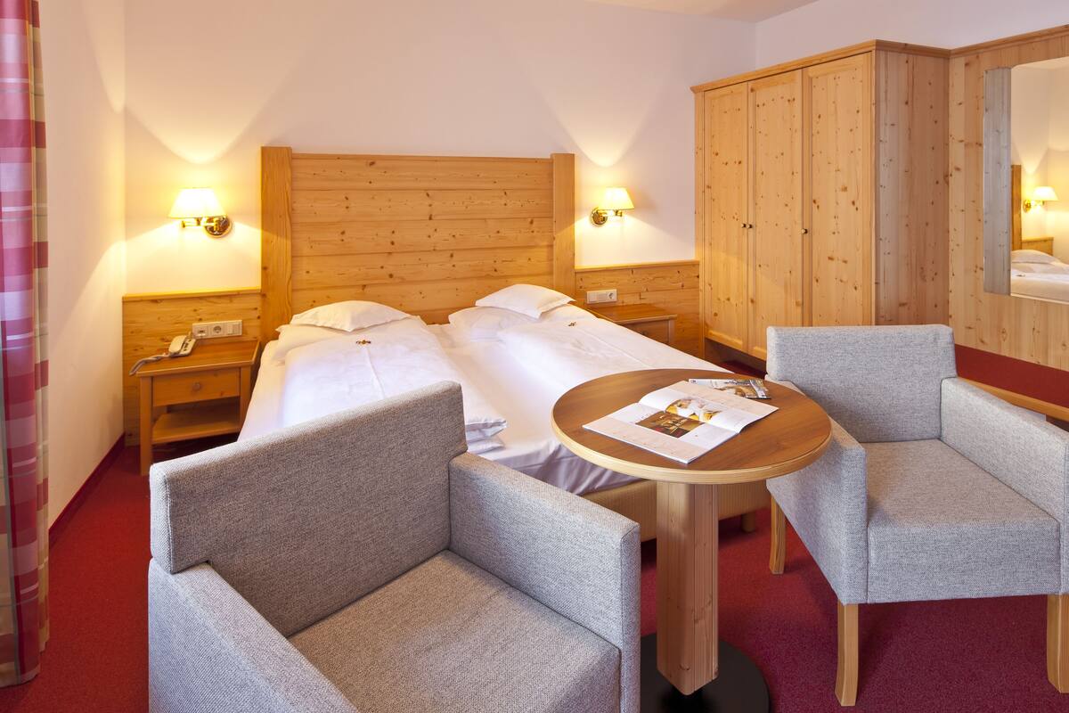 A hotel room with two single beds, a wooden wardrobe, a round table with a magazine, and two armchairs.