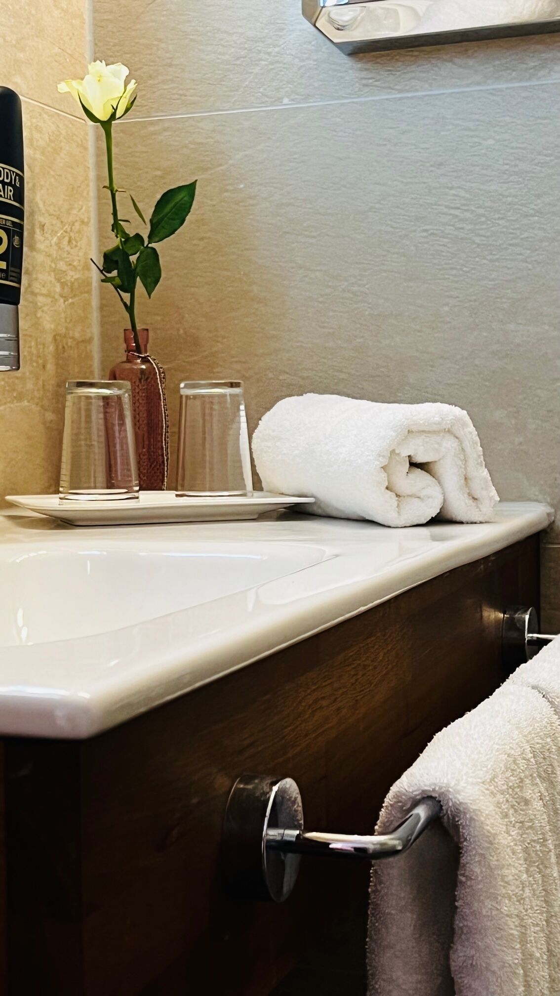 Un baño con un lavamanos blanco, una toalla, un florero de cristal con una flor y una botella de spray para el cuerpo y el ambiente.