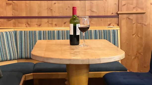 Een houten tafel met een fles wijn en een glas erop, geplaatst in een hoek met blauwe zachte zitplaatsen.