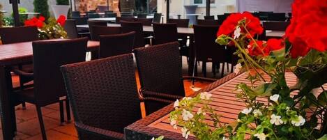 Un área de comedor al aire libre con mesas y sillas bajo sombrillas naranjas, rodeada de plantas en macetas y un muro de piedra.
