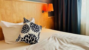 Een hotelkamer met een houten hoofdeinde, een wit bed met een kussen in wit en zwart patroon, een witte lamp en donkere gordijnen.