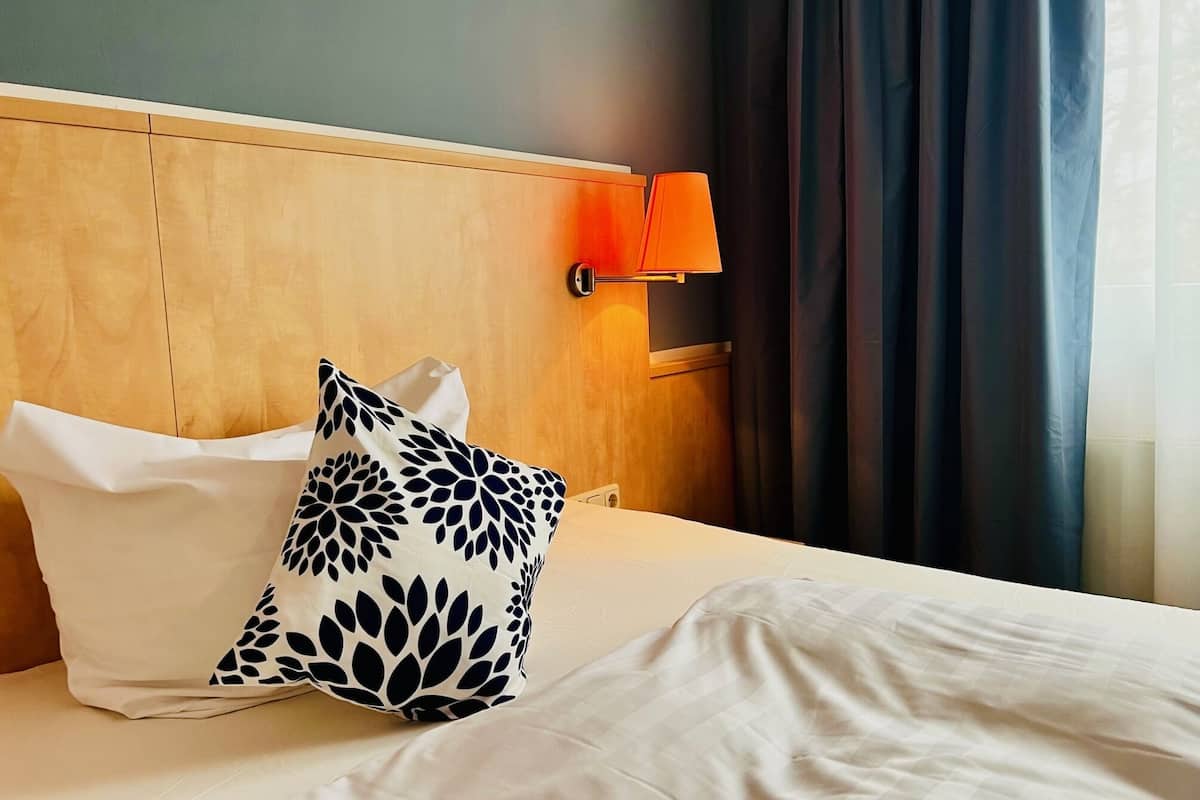 Ein Hotelzimmer mit einem Holzkopfteil, einem weißen Bett mit einem schwarz-weißen Kissenmuster, einer weißen Lampe und dunklen Vorhängen.