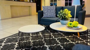 Een moderne lobby met een receptiebalie, een bank met decoratieve kussens, een ronde salontafel met een potplant en appels, en een vloerkleed met dessin.