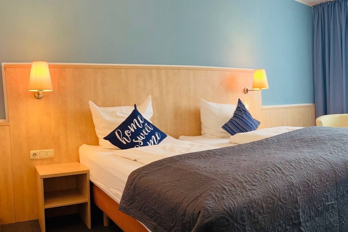 Ein Hotelzimmer mit Bett, Nachttisch, Sessel, Tisch mit einer Weinflasche und Snacks sowie einem Fenster mit Vorhängen.