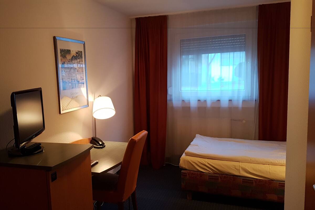 A hotel room with a bed, a desk with a computer, a chair, a lamp, a framed picture on the wall, and a window with curtains.