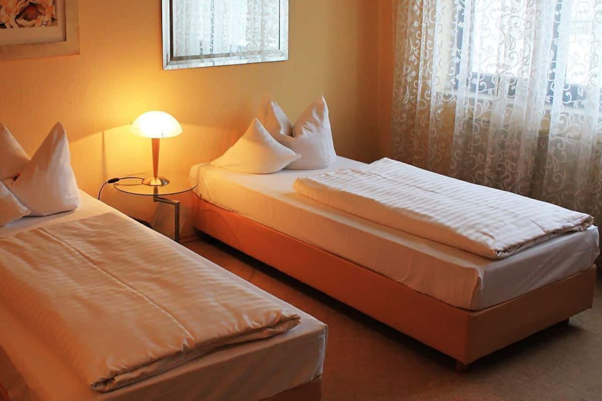 Dos camas individuales con ropa de cama y almohadas blancas, una mesita de noche con lámpara, y una ventana con cortinas translúcidas.