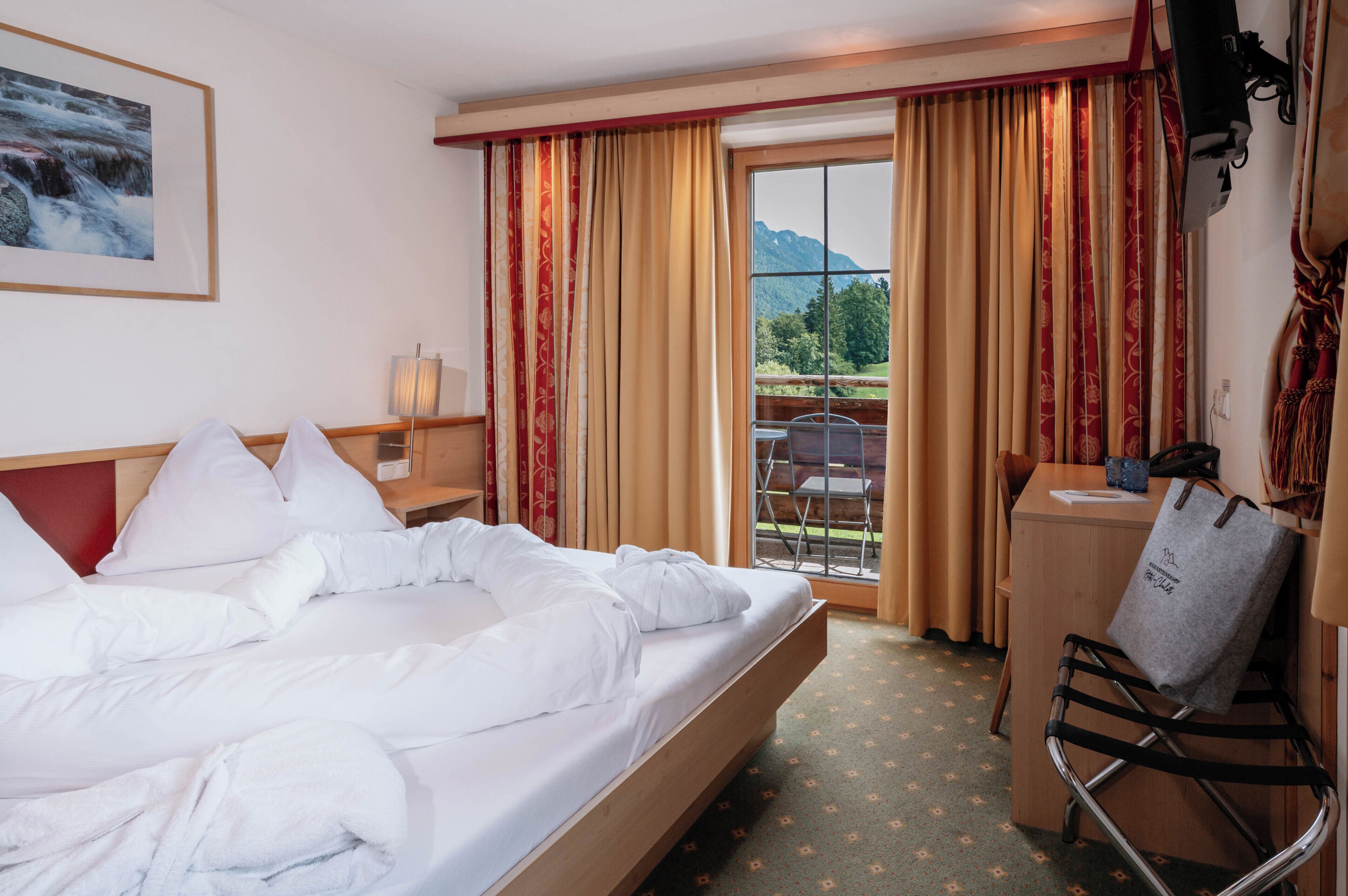 A hotel room with a bed, a desk, and a chair. There is a window with curtains and a view of the outdoors.