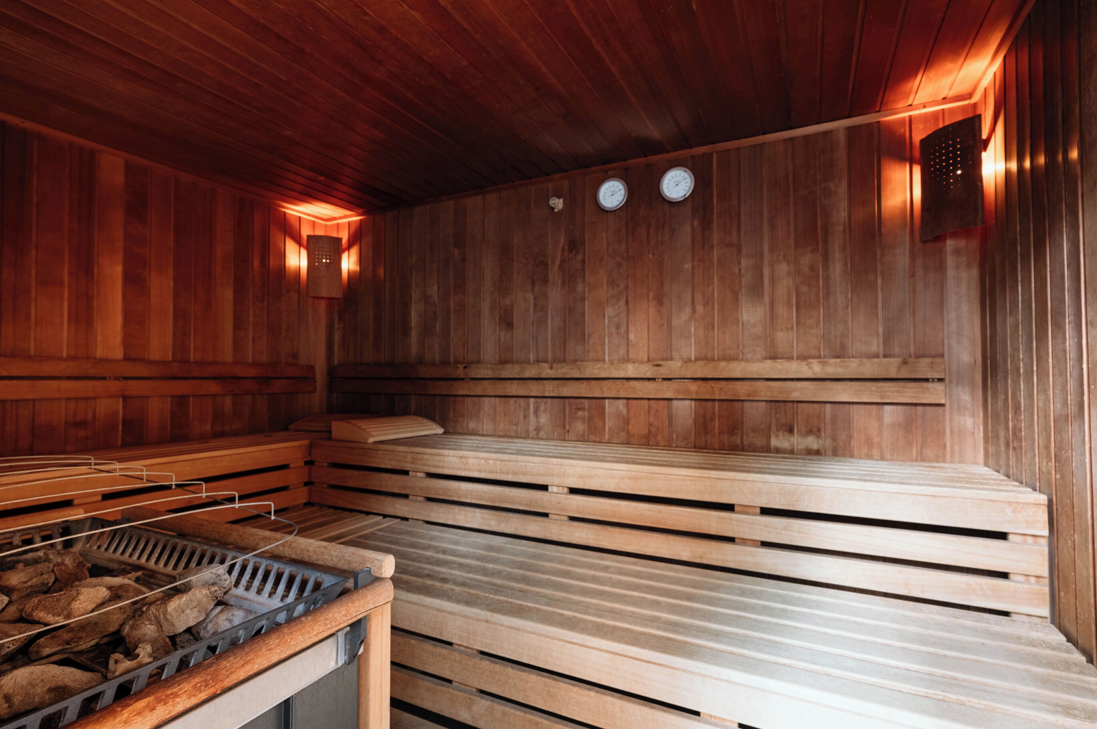 A sauna with wooden benches and a bench for sitting. There are two wall-mounted clocks and a heater with logs.
