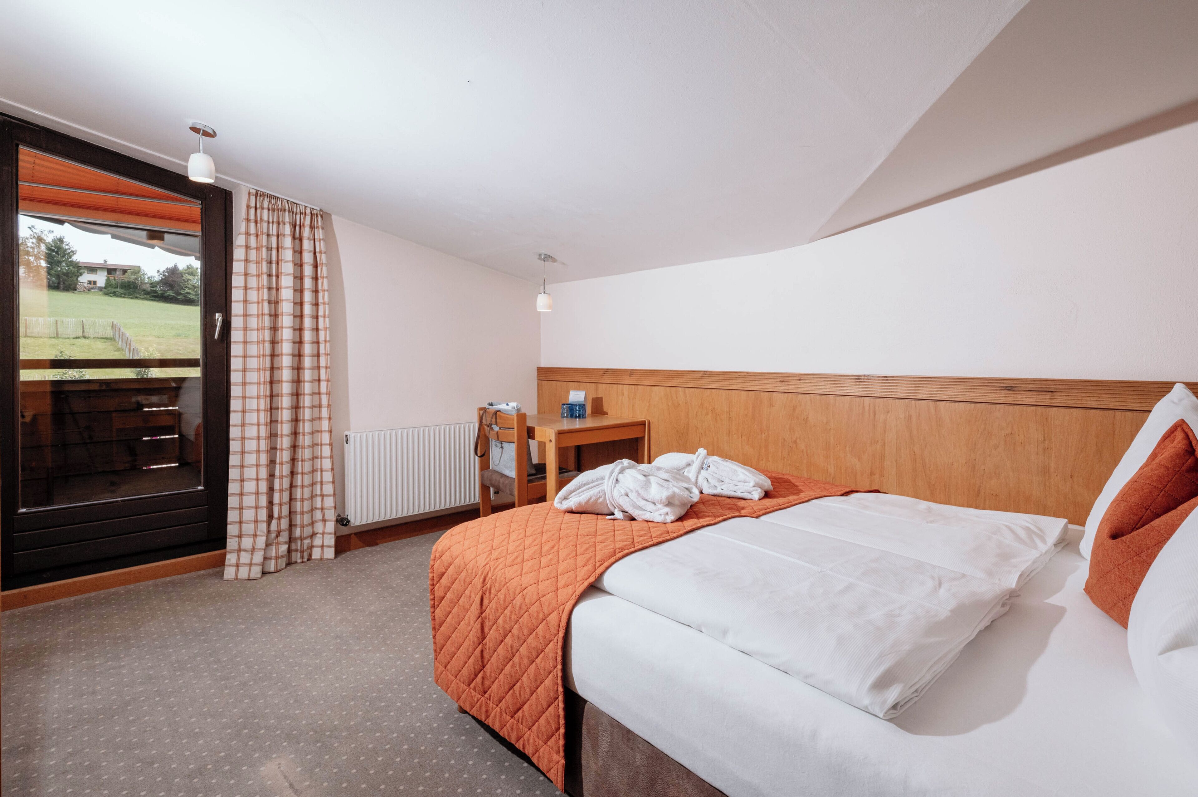 A hotel room with a bed, a wooden headboard, a window with a view of a grassy area, and a radiator.