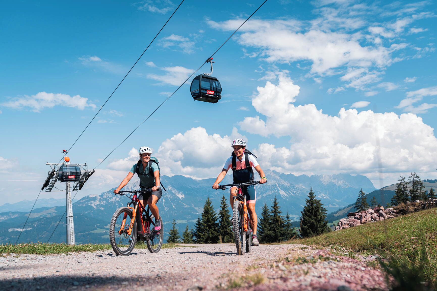 Two cyclists on a mountain road with a cable car in the background.
