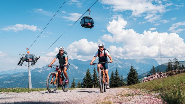 Dos ciclistas en una carretera de montaña, con un teleférico al fondo.