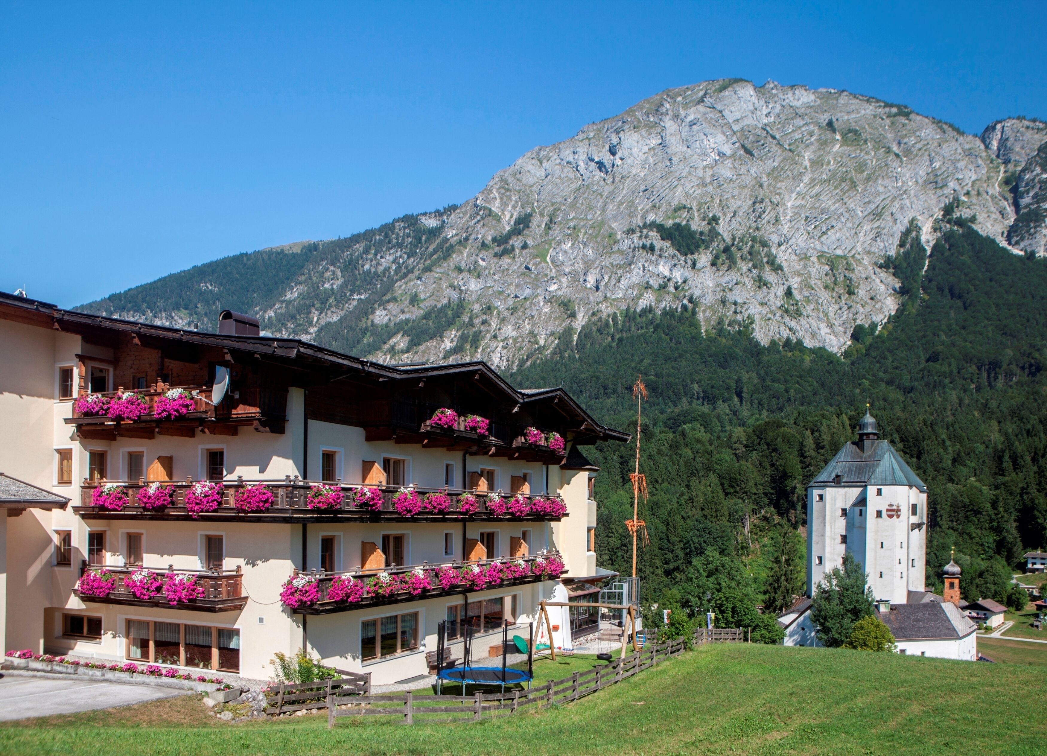 Un hotel con balcones decorados con flores, un paisaje montañoso de fondo y una iglesia pequeña cerca.