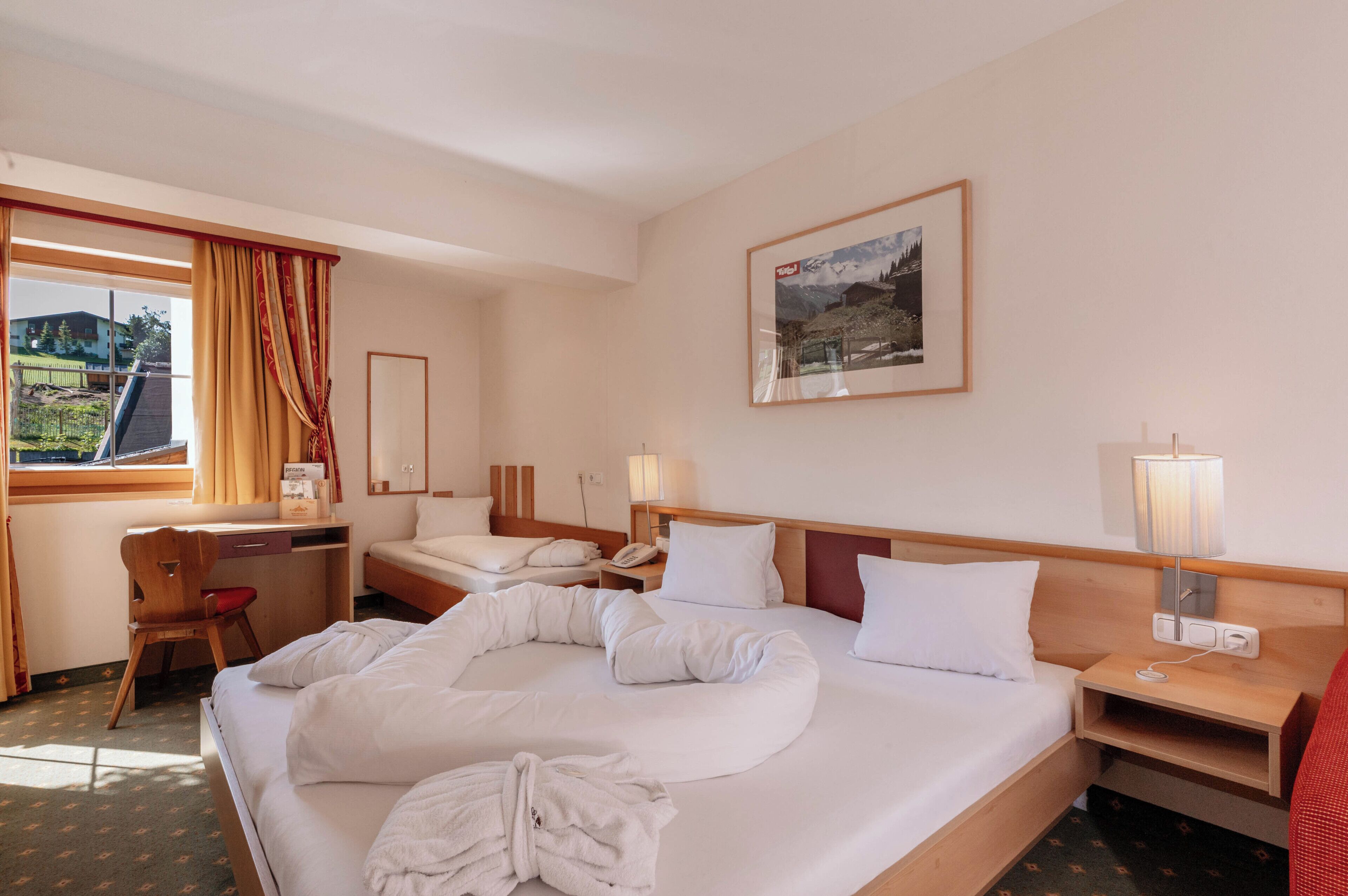 A hotel room with a bed, a desk, a chair, a bedside table with a lamp, and a window with curtains.