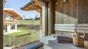 A wooden spa room with a bench, a wooden bucket, and a view of a house and grassy area outside. - Hotel Mariasteiner Hof (Mariastein)