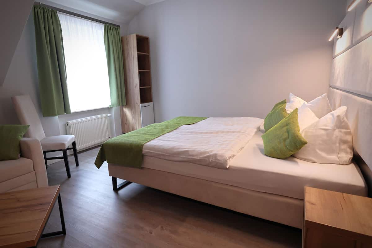 Un dormitorio con cama, una silla, una mesita, un estante y una ventana con cortinas.