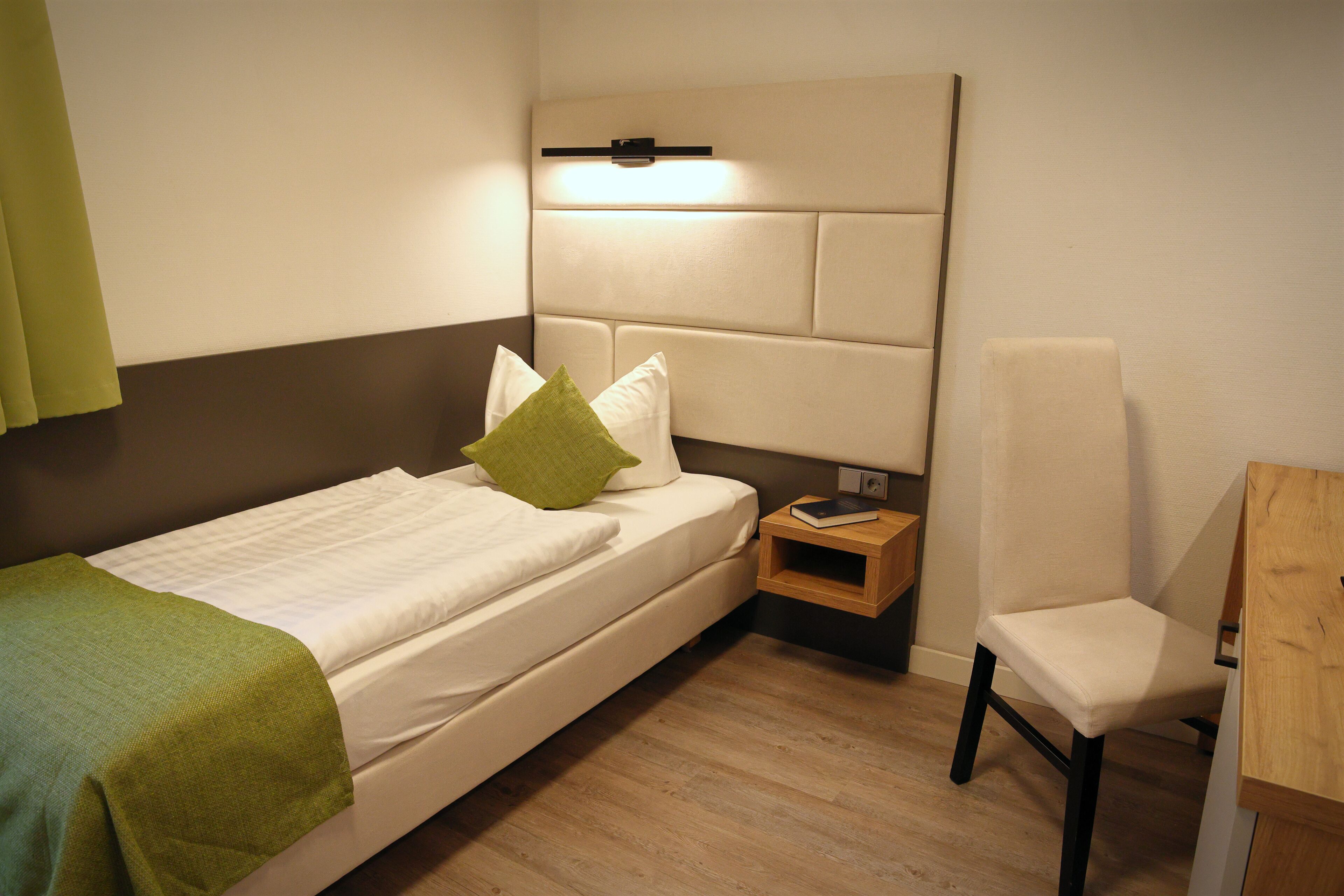 A neatly made bed with a green pillow, a bedside table with a book, and a chair in a modern room with wooden flooring and a headboard with built-in lighting.