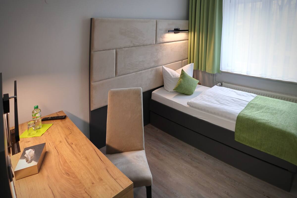 Habitación de hotel con escritorio de madera, una silla, una cama con almohada verde y cabecero.