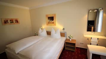 Ein Hotelzimmer mit einem großen Bett, einem Nachttisch mit Lampe, einem kleinen Schreibtisch, einem Stuhl, einem Spiegel und Bildern an den Wänden.