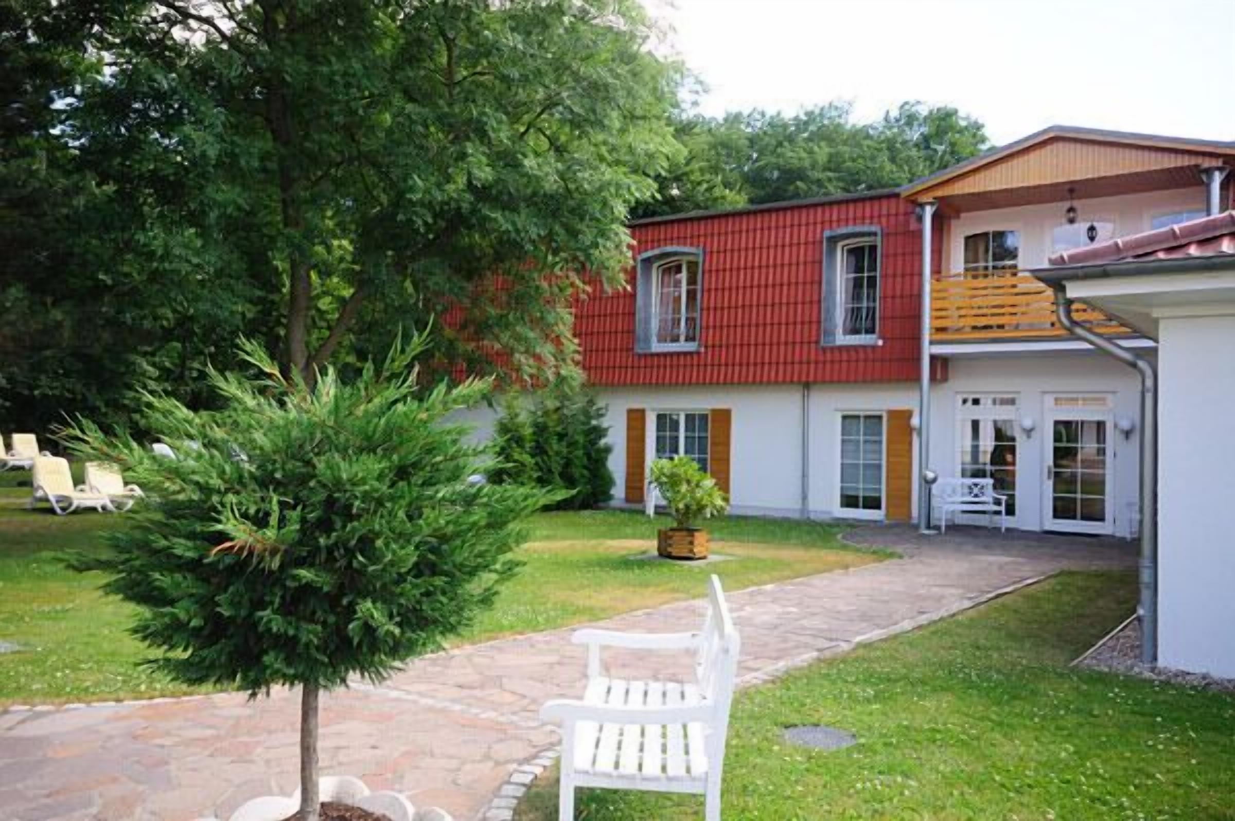 Ein zweigeschossiges Haus mit roter Fassade, einer weißen Haustür und einem kleinen Vorgarten mit Bank und Baum.