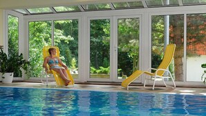 Una piscina interior con paredes de vidrio transparente, dos sillones de descanso y una planta en maceta.