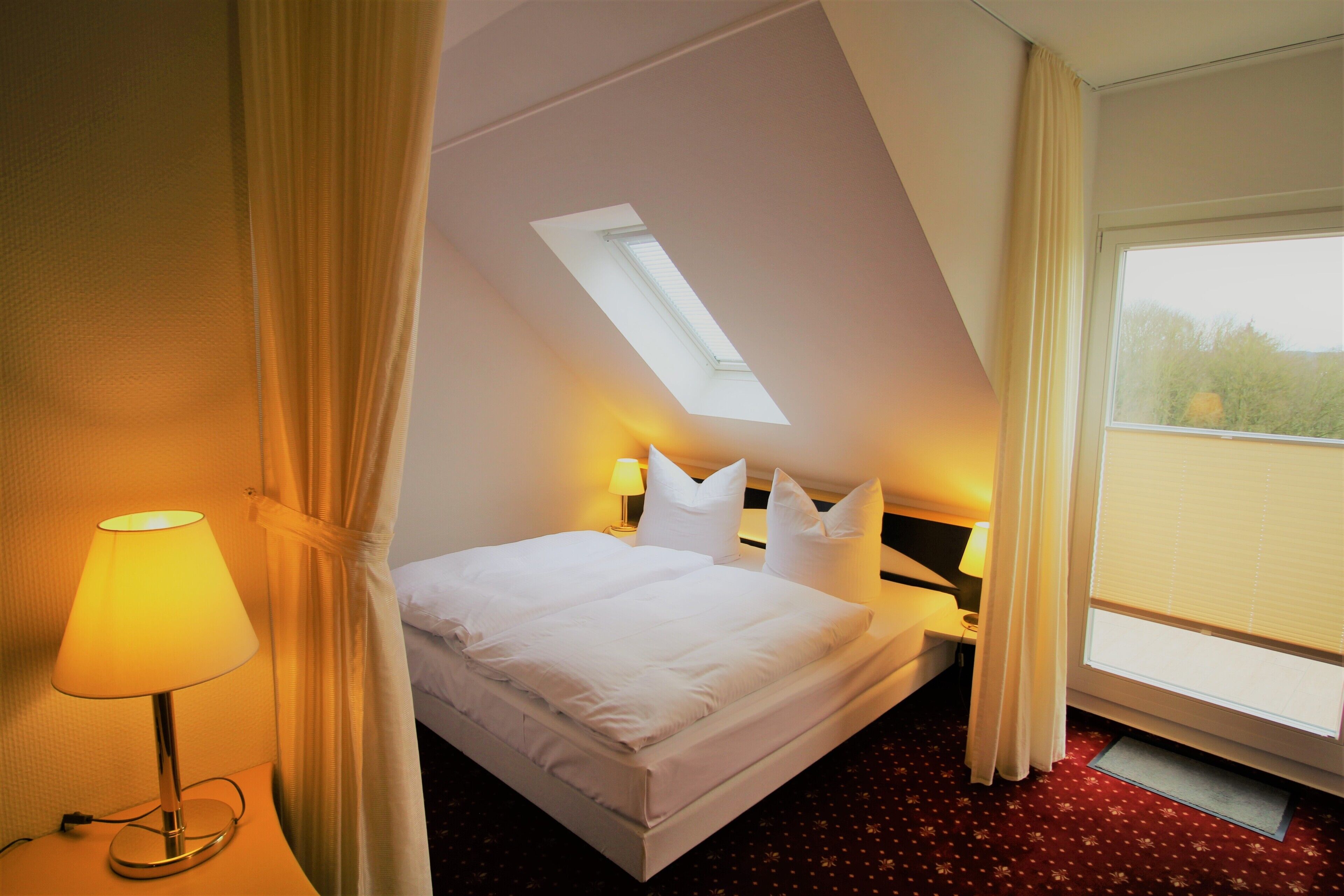 Ein Hotelzimmer mit einem Bett, einem Nachttisch, einer Lampe, einem Fenster mit Vorhängen und einem Oberlicht.