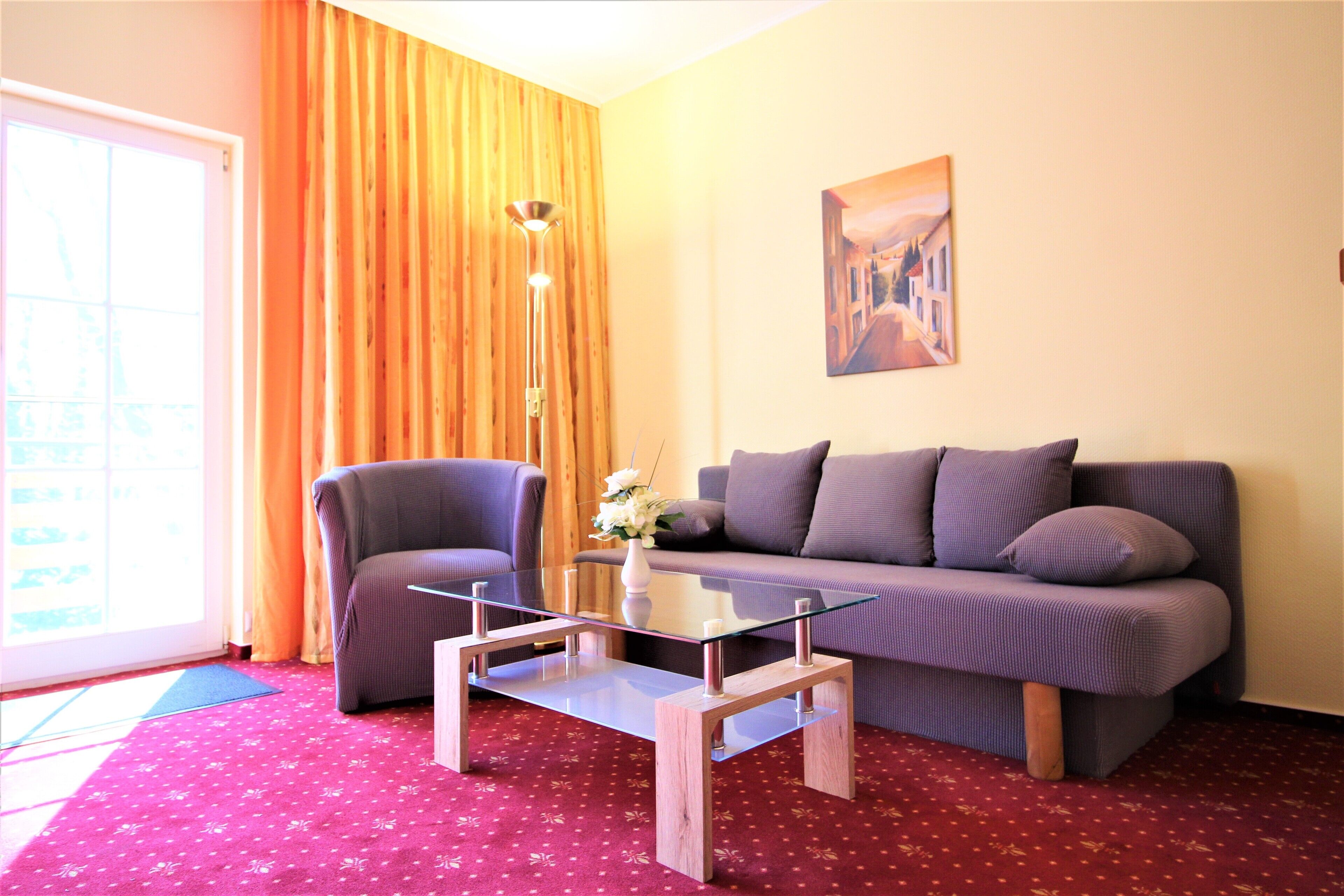 A living room with a glass coffee table, a sofa, a chair, and a painting on the wall.
