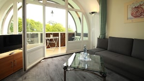 Una sala moderna con una mesa de centro de vidrio, un sofá gris y un televisor sobre un mueble de madera. Un amplio ventanal arqueado ofrece vistas al exterior.