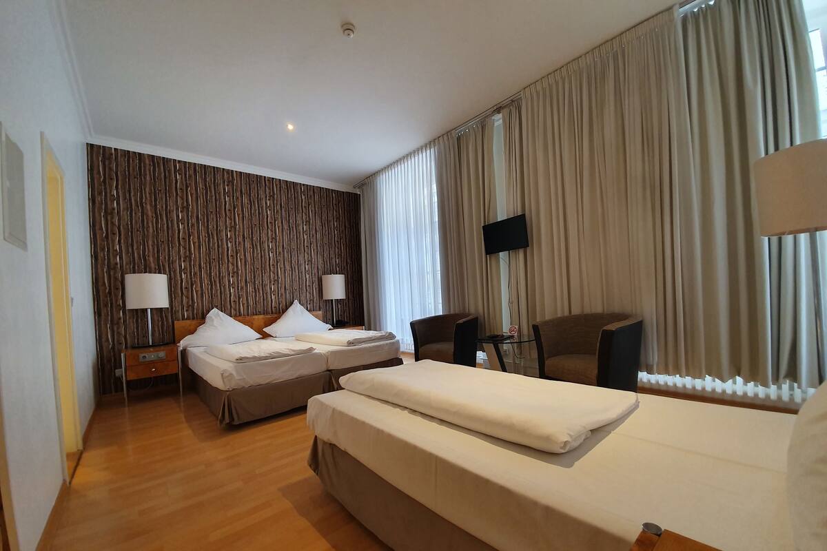 A hotel room with two beds, a wooden accent wall, a flat-screen TV, and a large window with curtains.