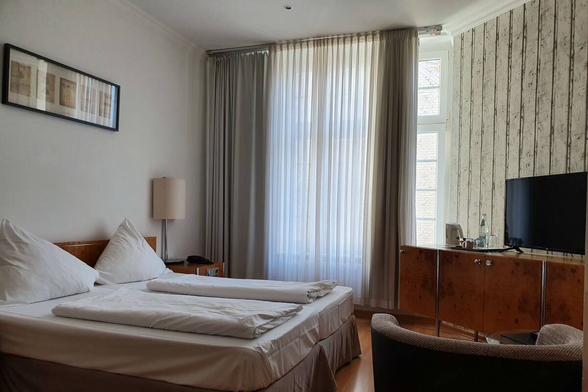 A hotel room with two beds, a desk with a TV, a chair, and a large window with curtains.