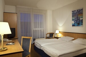 Quarto de hotel com cama, escrivaninha com luminária, cadeira, televisão e um quadro na parede.